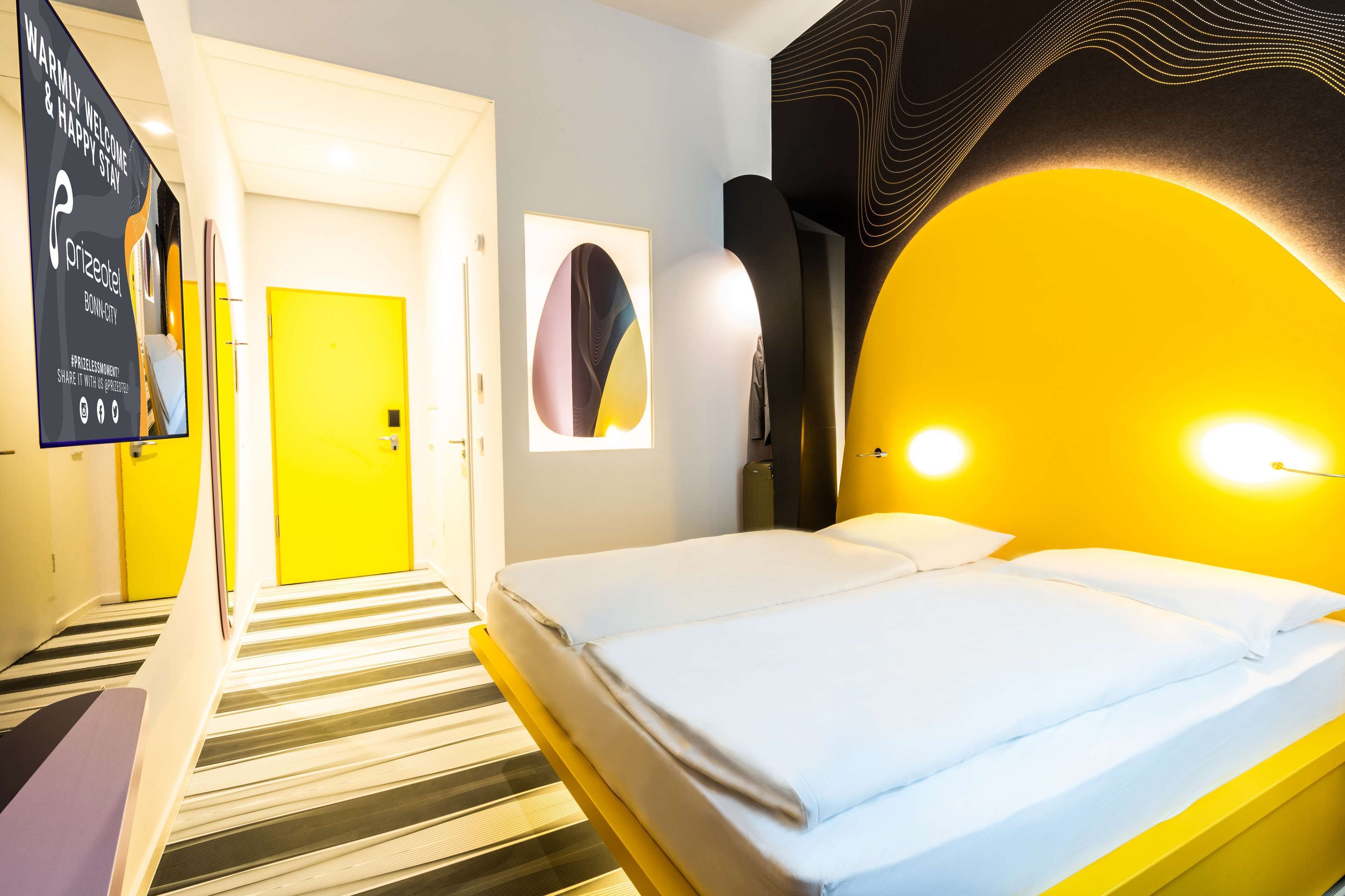 A hotel room with a bed, a yellow accent wall, a framed abstract artwork, and a digital screen displaying a welcome message.