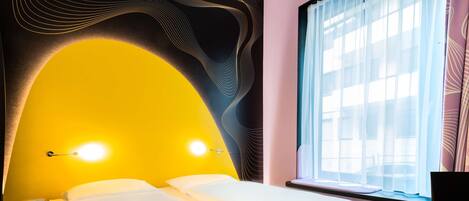 A hotel room with a bed, bedside lamps, a window with sheer curtains, and a television.