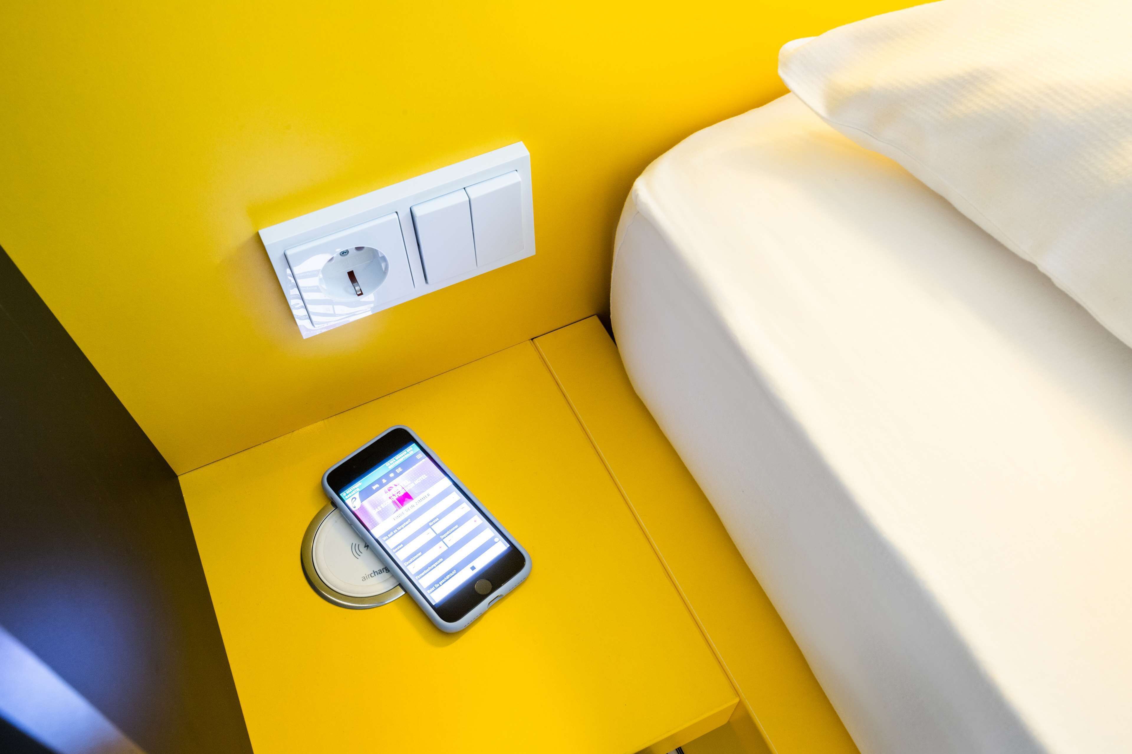 A smartphone on a yellow bedside table with a white bed and a wall-mounted switch.