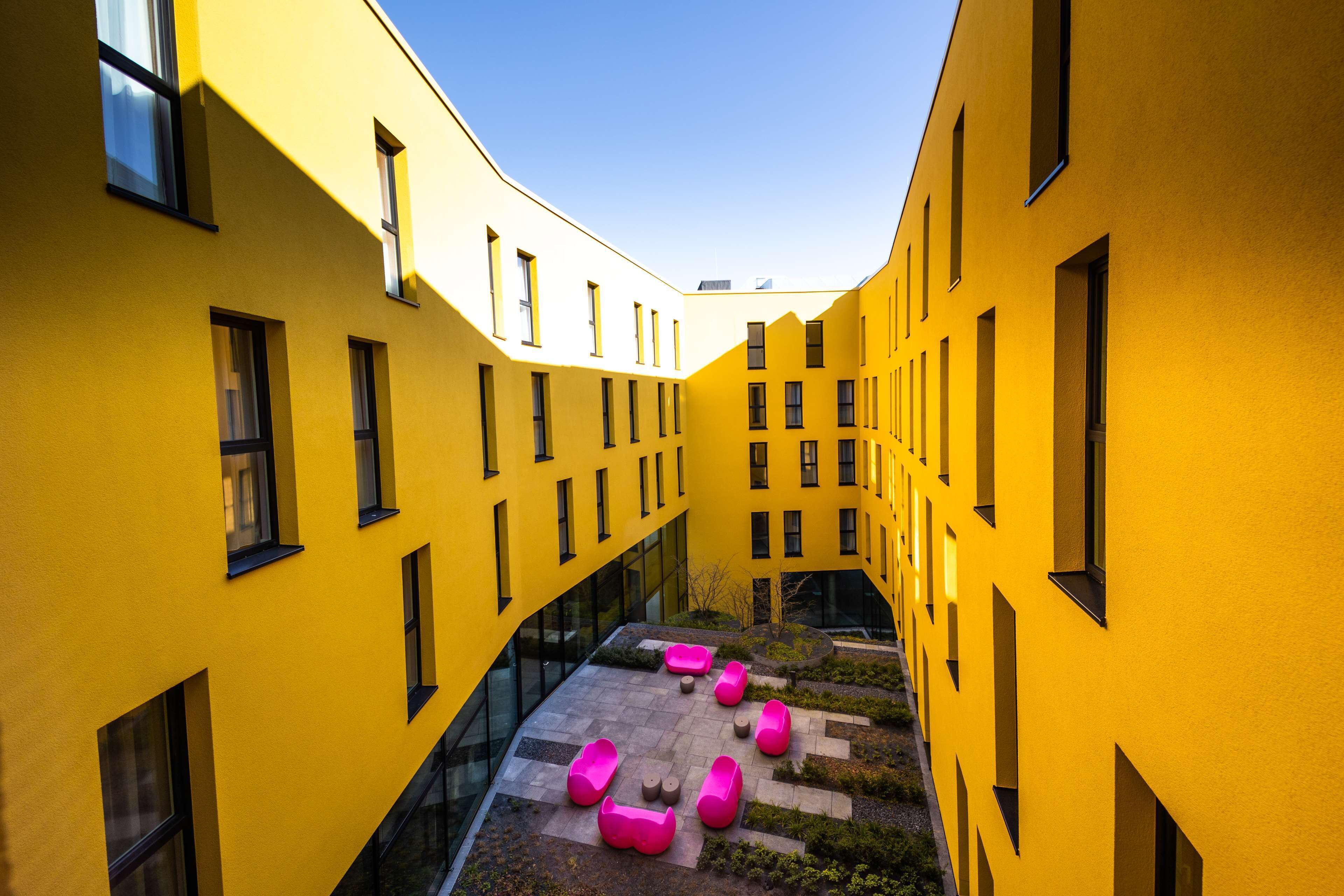 Ein gelbes Gebäude mit vielen Fenstern und einem Innenhof mit rosa Sitzgelegenheiten.