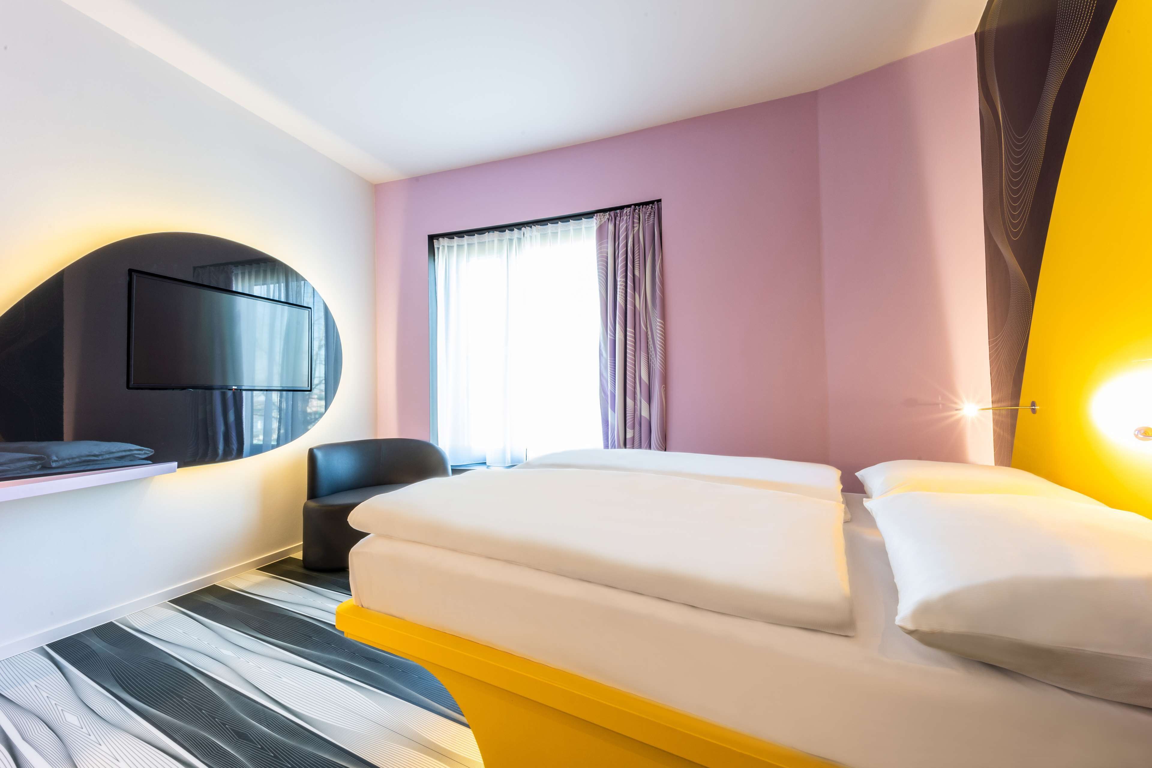 Hotellihuone, jossa on sänky, tuoli, seinälle asennettu televisio ja ikkuna, jossa on verhot.