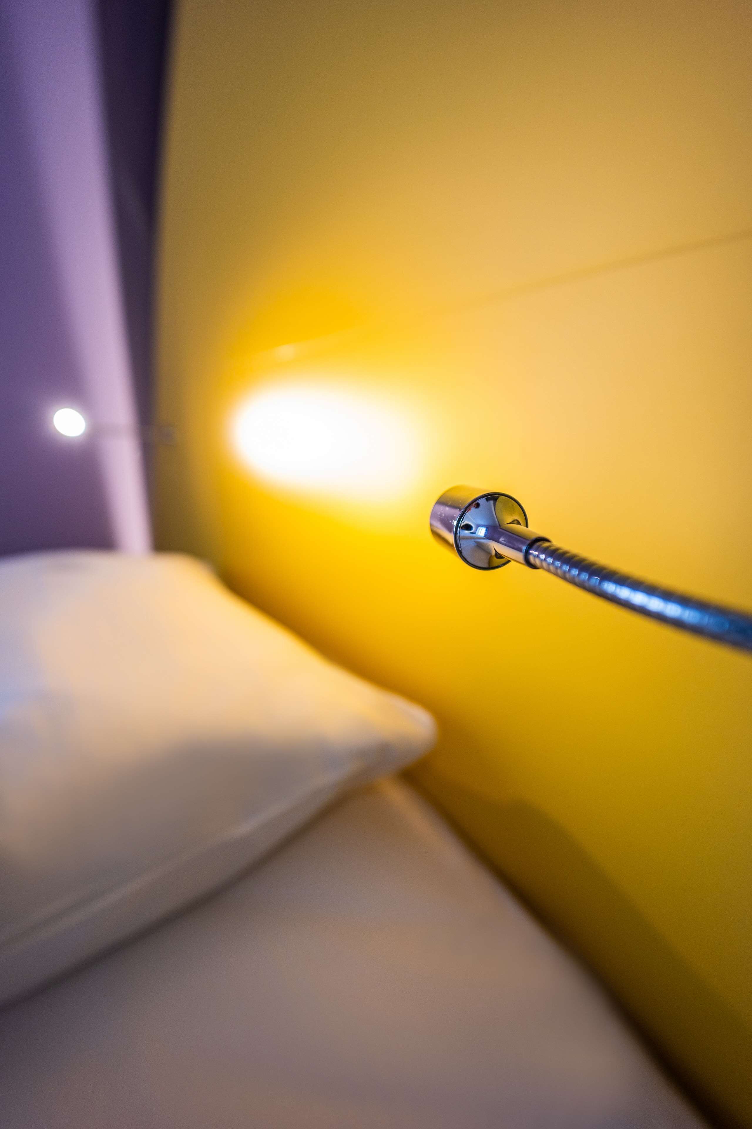 A close-up of a metallic object, possibly a part of a bed or a piece of furniture, against a yellow wall with a soft light source nearby.