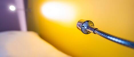 A close-up of a metallic object, possibly a part of a bed or a piece of furniture, against a yellow wall with a soft light source nearby.