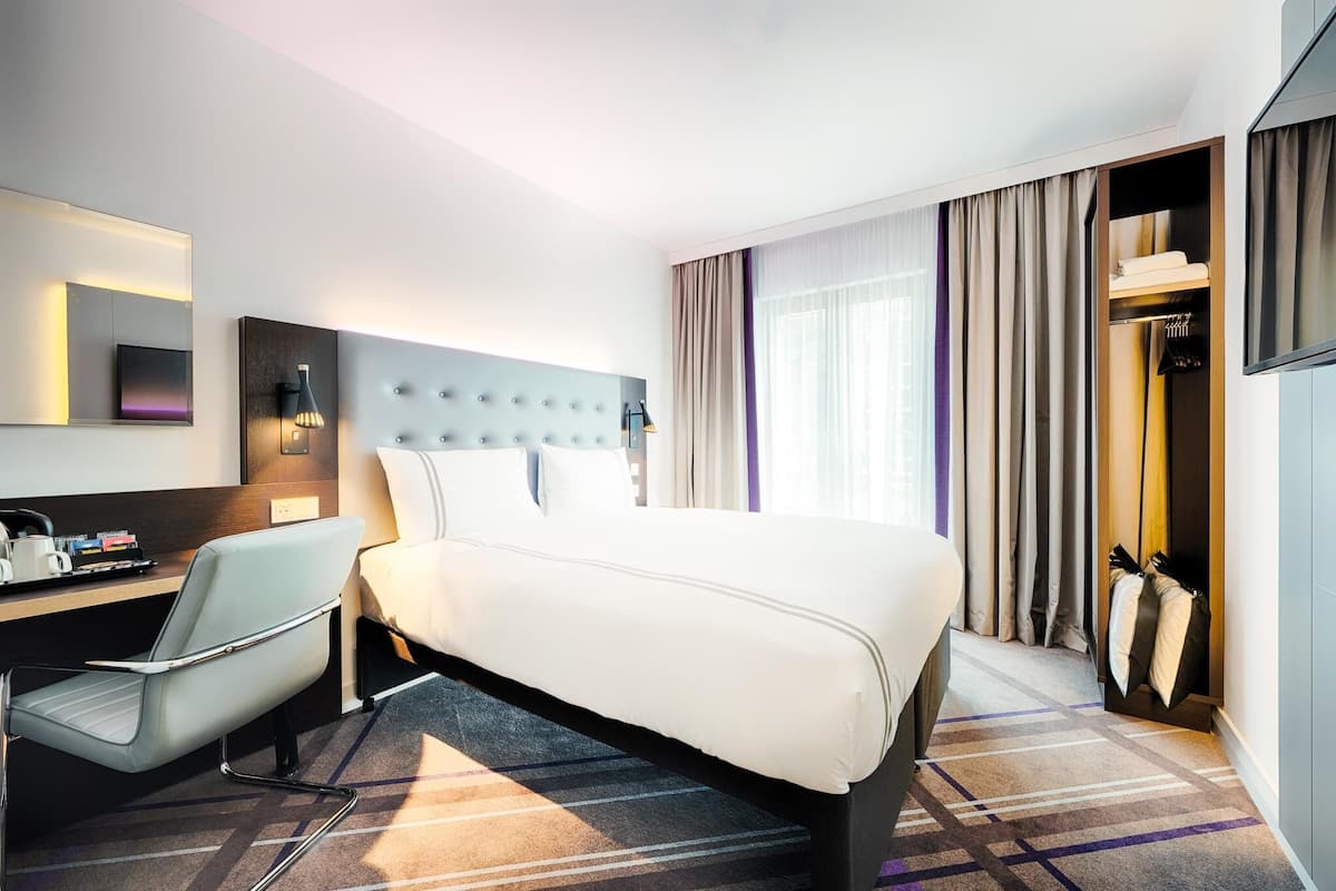 A hotel room with a bed, a desk, a chair, a wardrobe, and a large window with curtains.