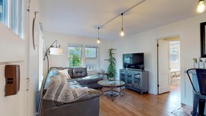 Una sala de estar moderna con un sofá de cuero, una mesa de centro de vidrio, un televisor sobre un mueble bajo y un ventanal con persianas.