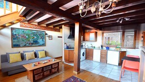 A spacious living area with a sofa, coffee table, and a colorful rug. A kitchen with wooden cabinets and a washing machine is visible in the background.