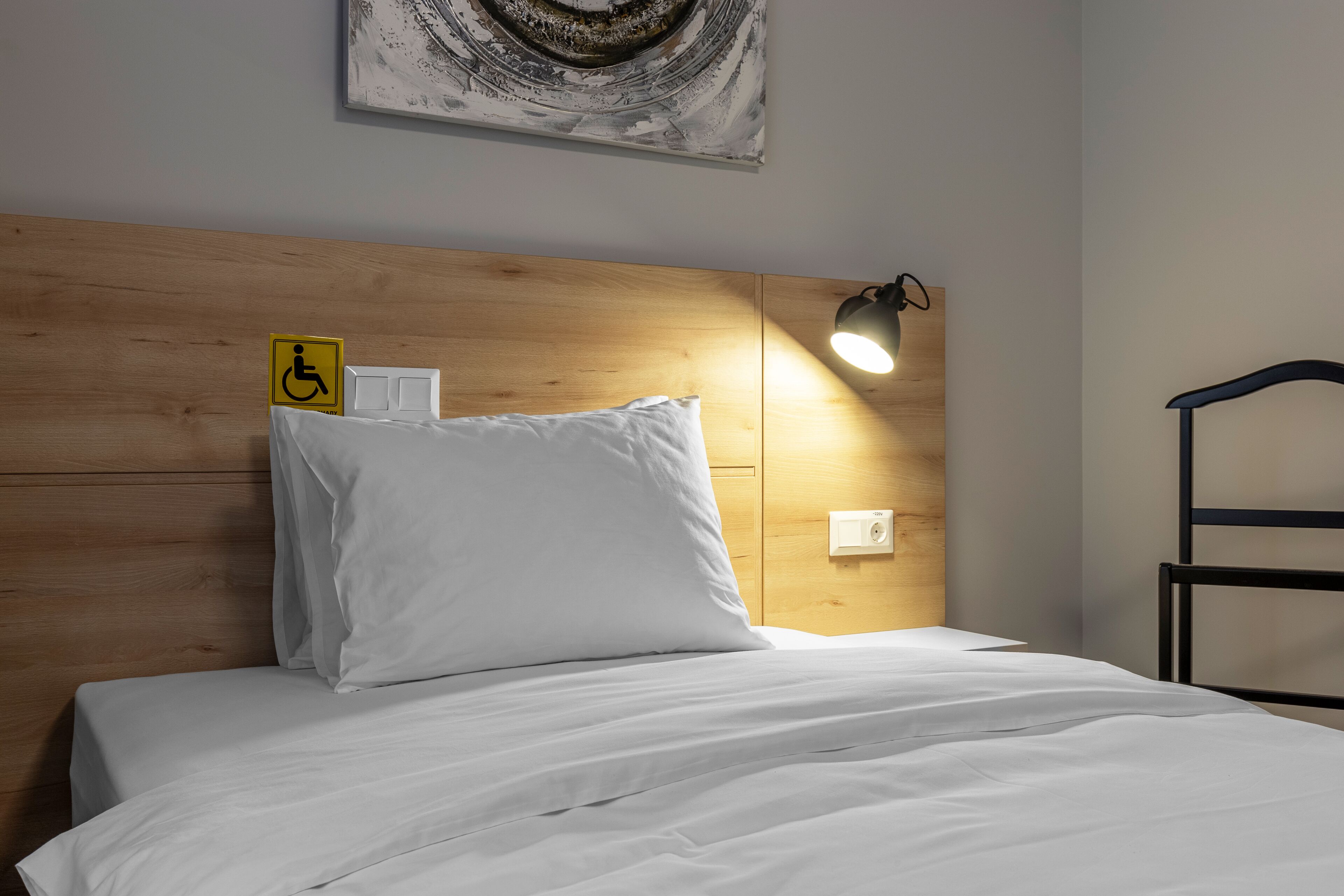 A neatly made bed with white linens, a wooden headboard, a bedside lamp, and a wheelchair accessibility sign.