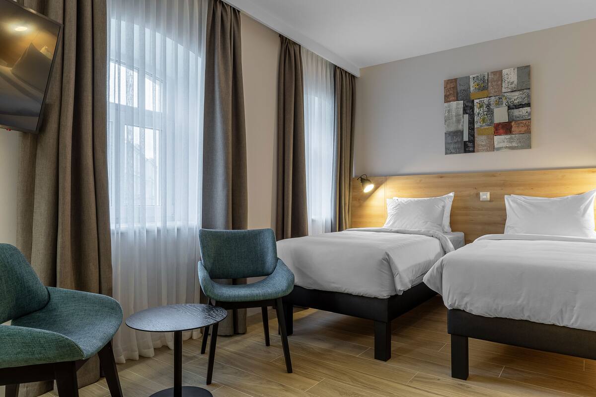 A hotel room with two beds, a chair, a small table, and a wall-mounted artwork.