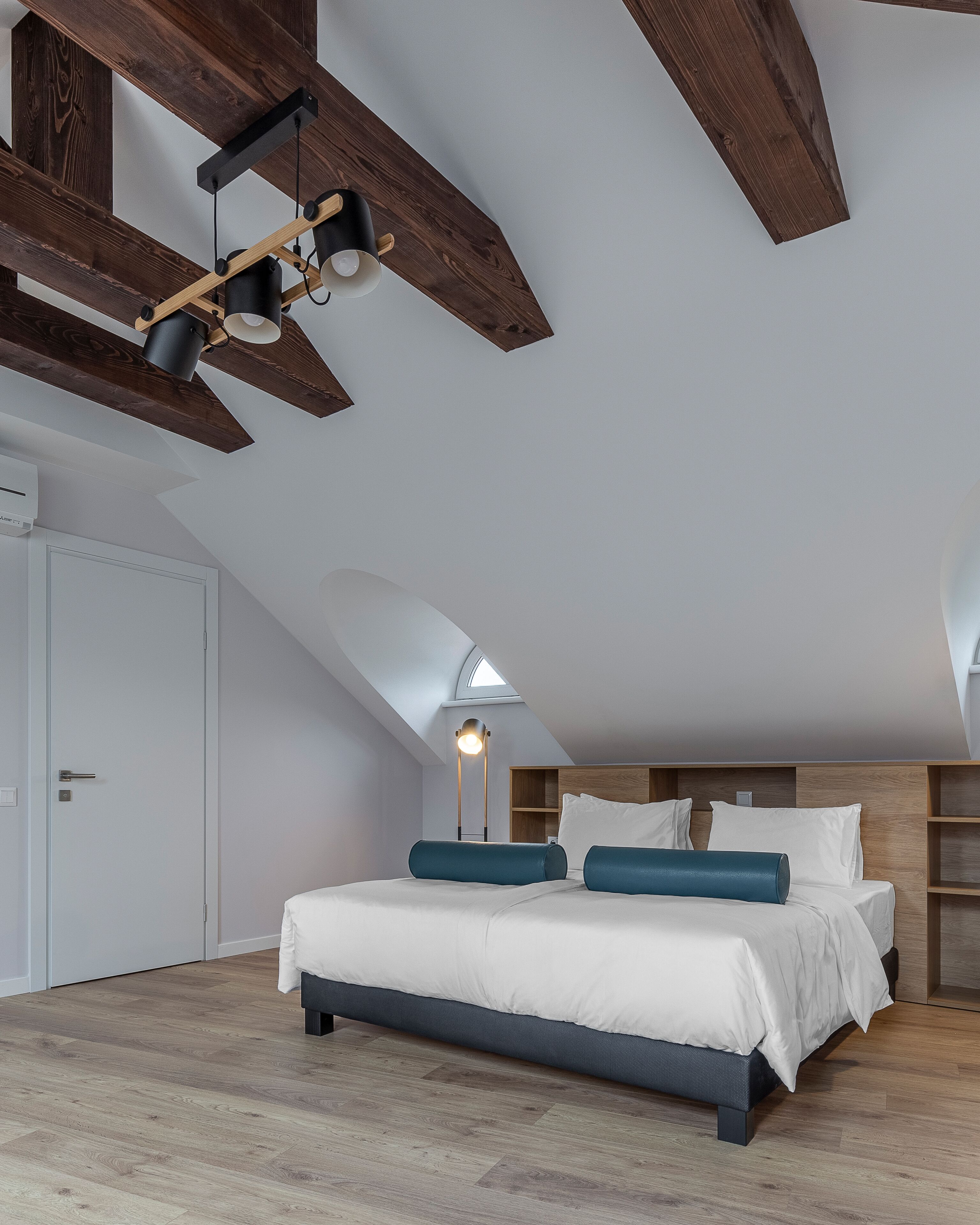 A modern bedroom with a sloped ceiling, a skylight, a bed with white bedding and blue pillows, and a floor lamp.
