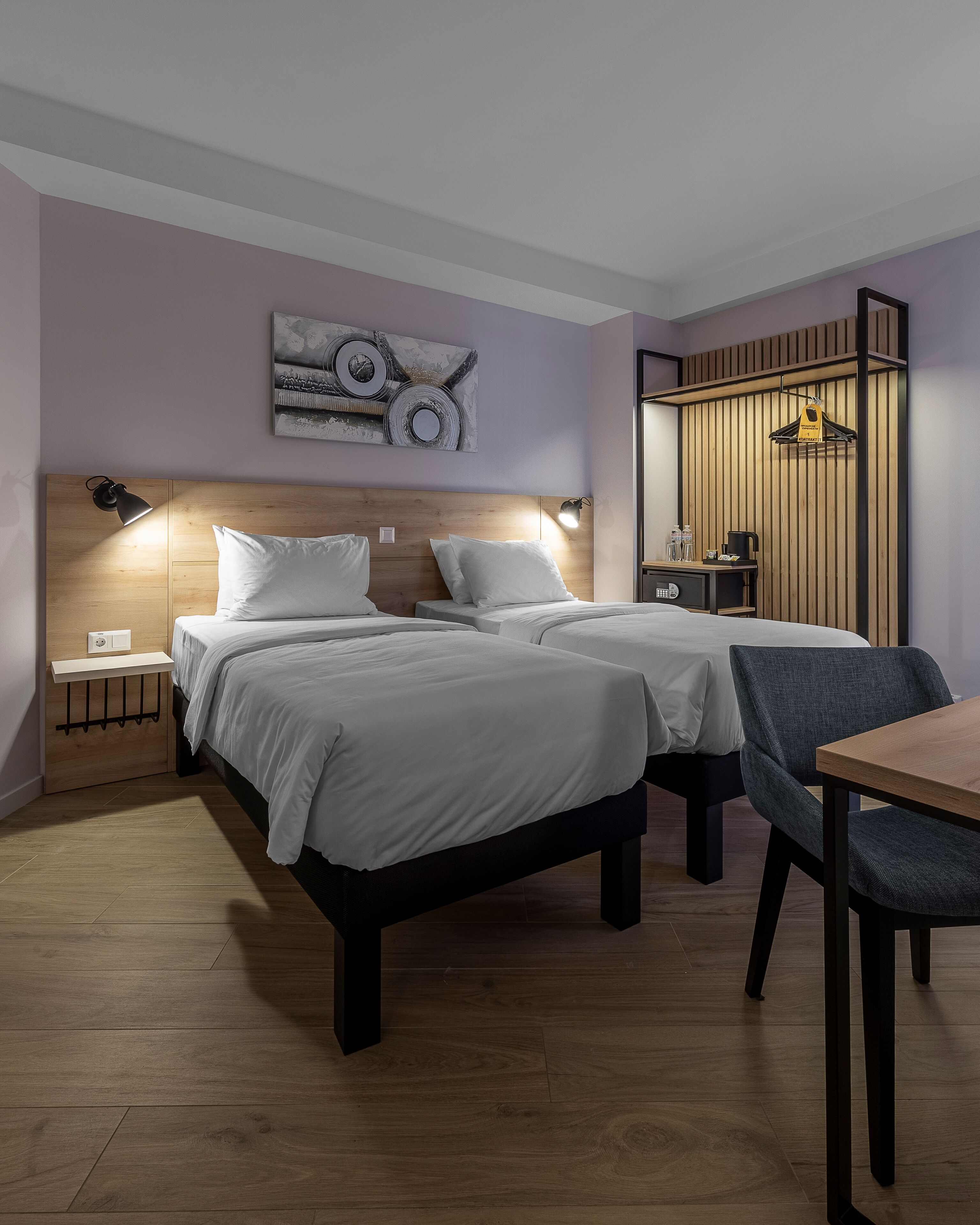 Quarto de hotel com duas camas, uma escrivaninha e uma cadeira. Há uma luminária na parede e um quadro sobre as camas.