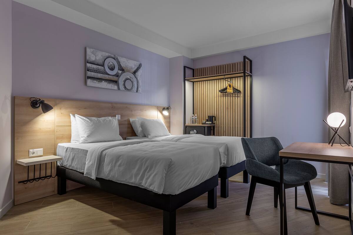 A modern bedroom with a wooden headboard, a bed with white bedding, a bedside table with a lamp, a chair, and a small table with a lamp.