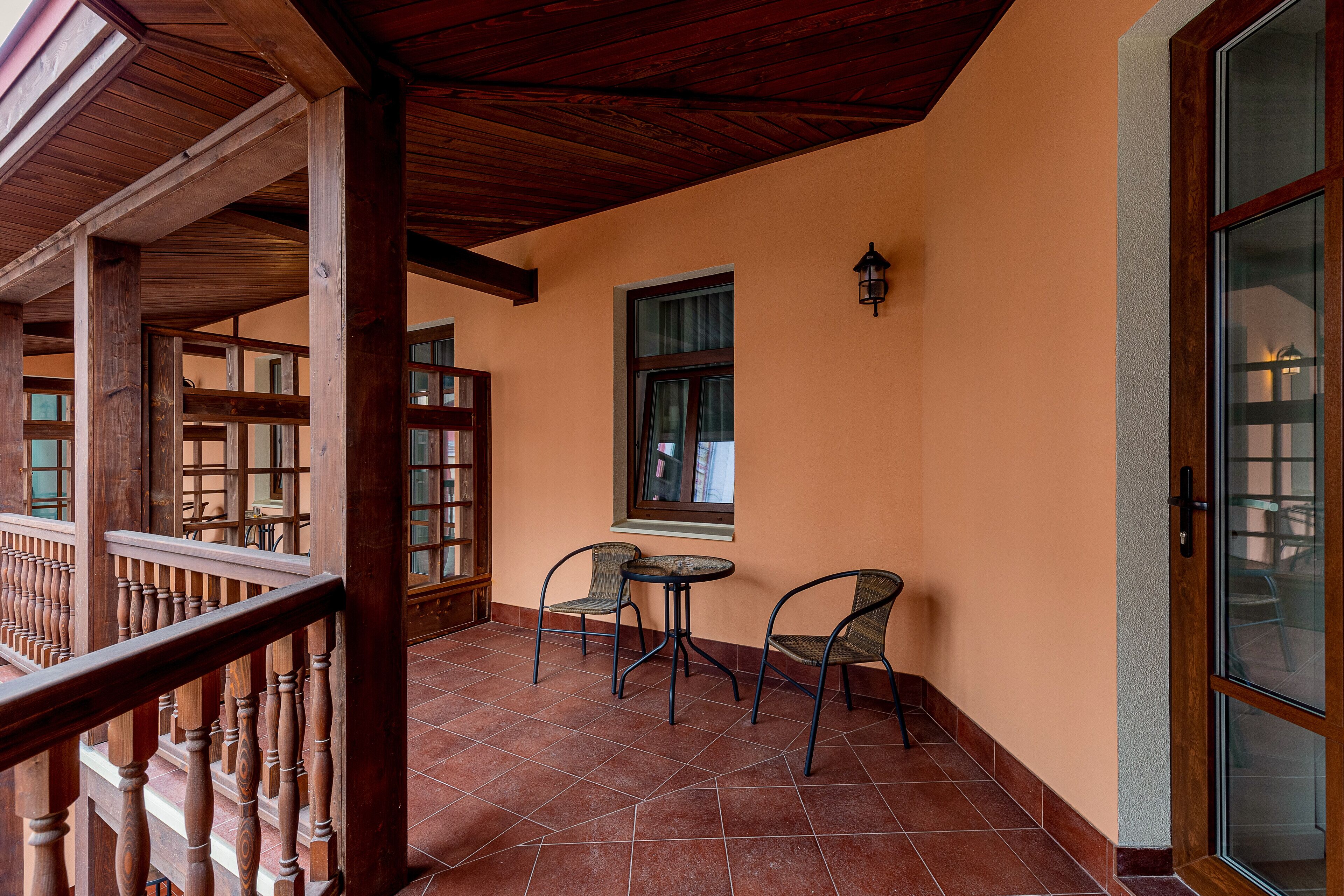A balcony with wooden railings and a tiled floor, a small table with two chairs, and a window with a curtain.