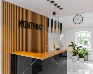 Ein Empfangsschalter mit einer Wand aus Holzleisten und dem Schriftzug "KONTRAKT" in schwarzen Buchstaben. Über dem Schalter hängen Pendelleuchten, an der Wand hängt eine Uhr und rechts befinden sich Blumentöpfe.