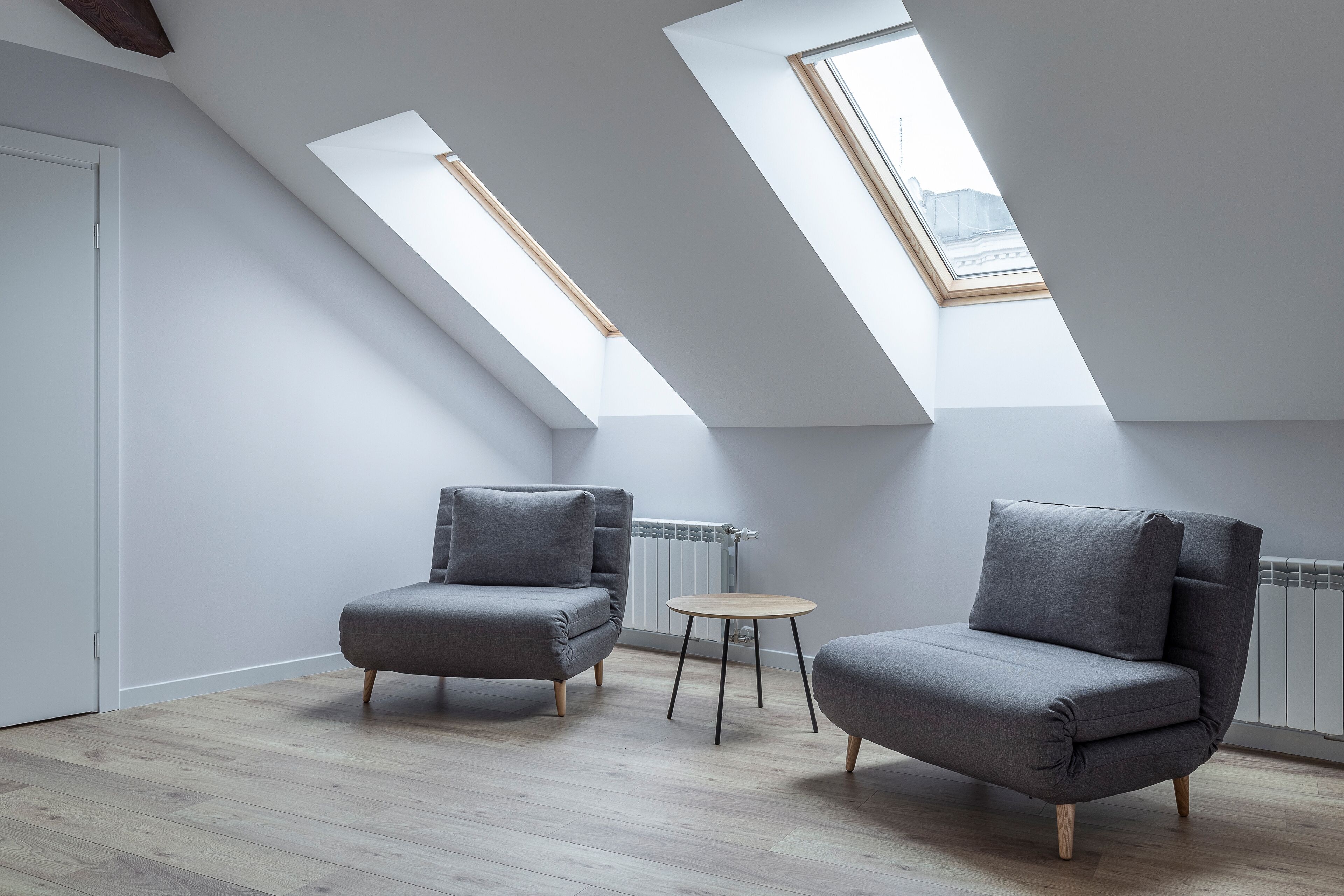 A modern loft bedroom with skylights, a grey sofa, a small round table, and a radiator.