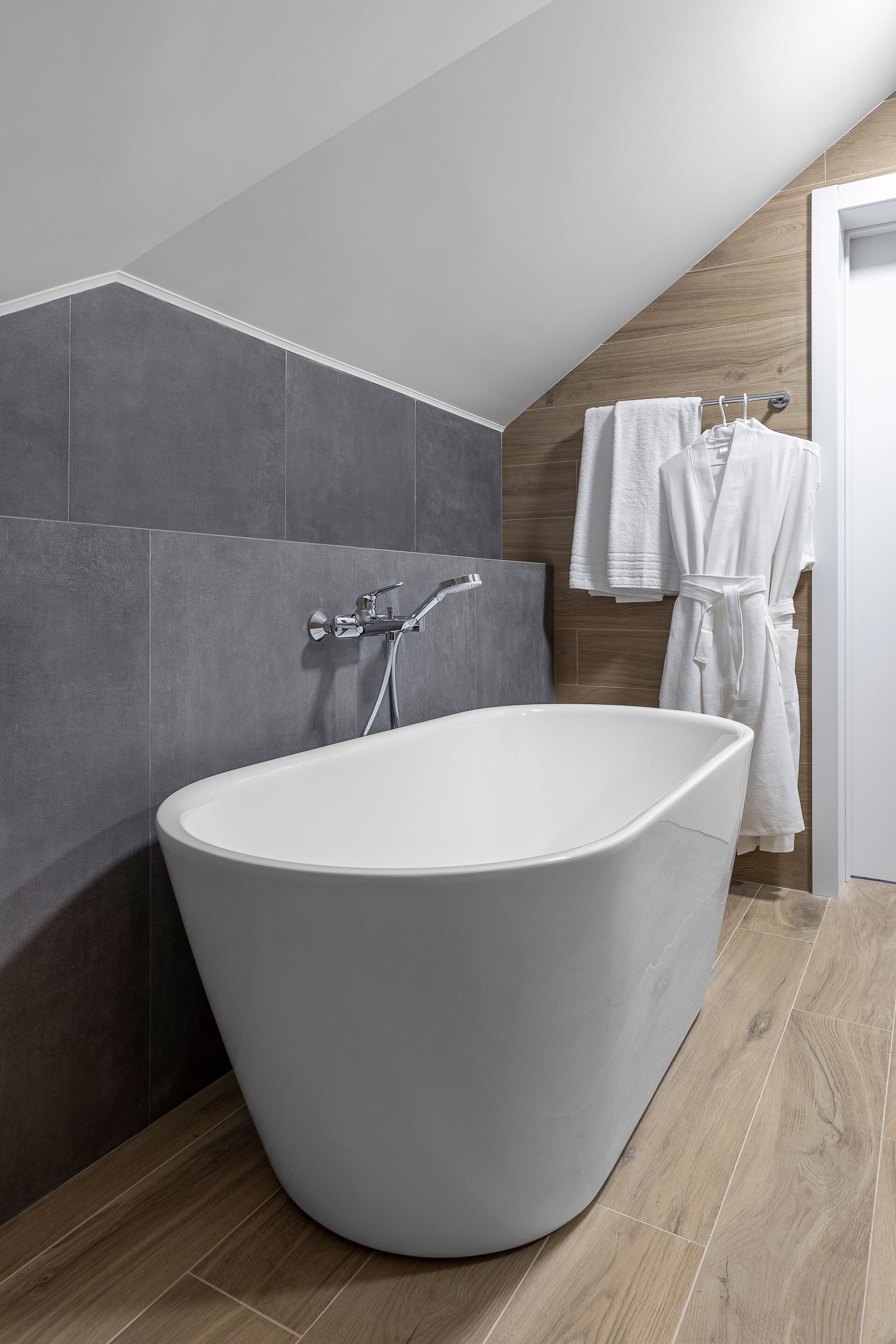 A modern bathroom with a freestanding bathtub, grey tiles, wooden accents, and hanging towels.