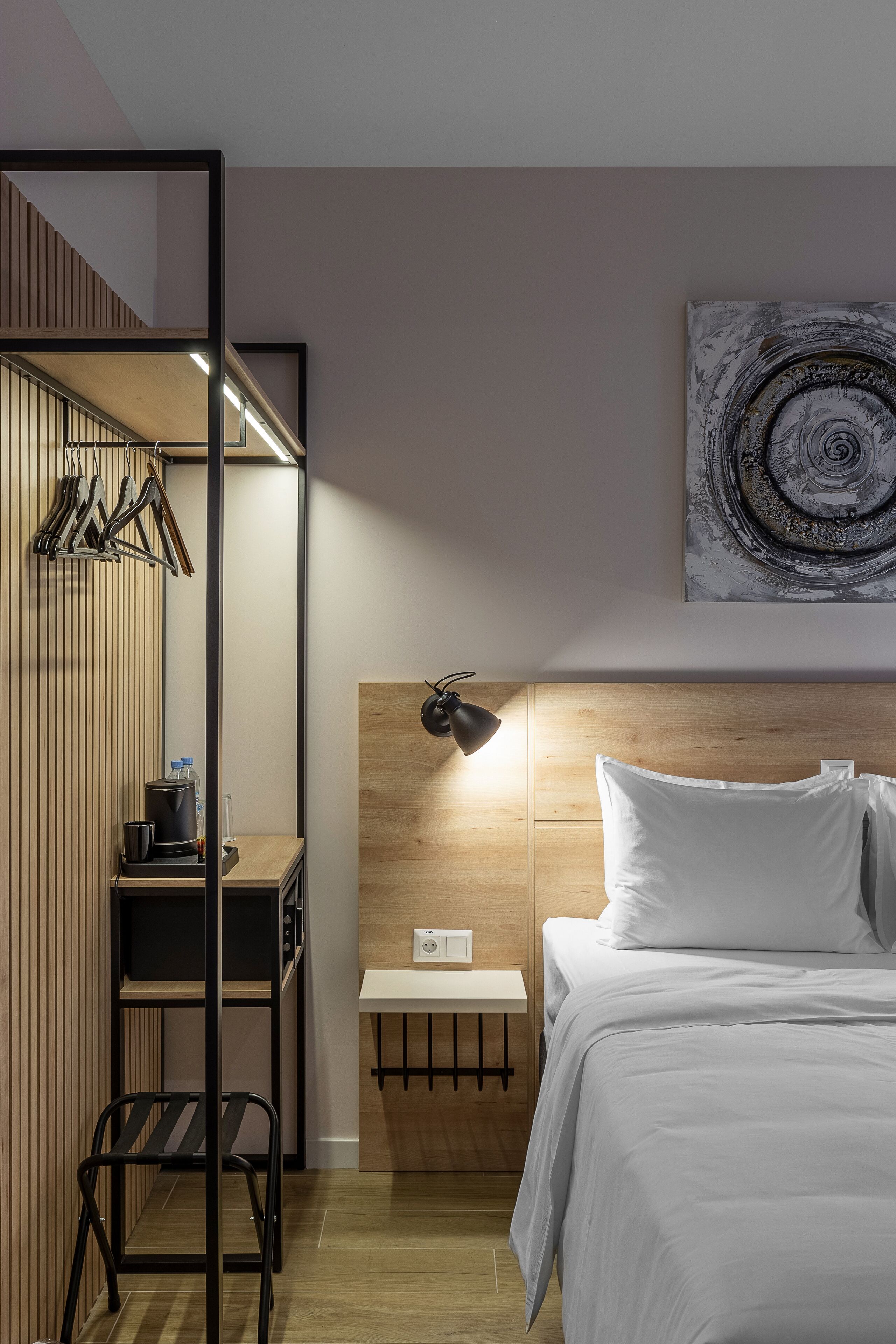 A modern hotel room with a wooden headboard, a bed with white bedding, a desk with a lamp, and a wall-mounted abstract art piece.