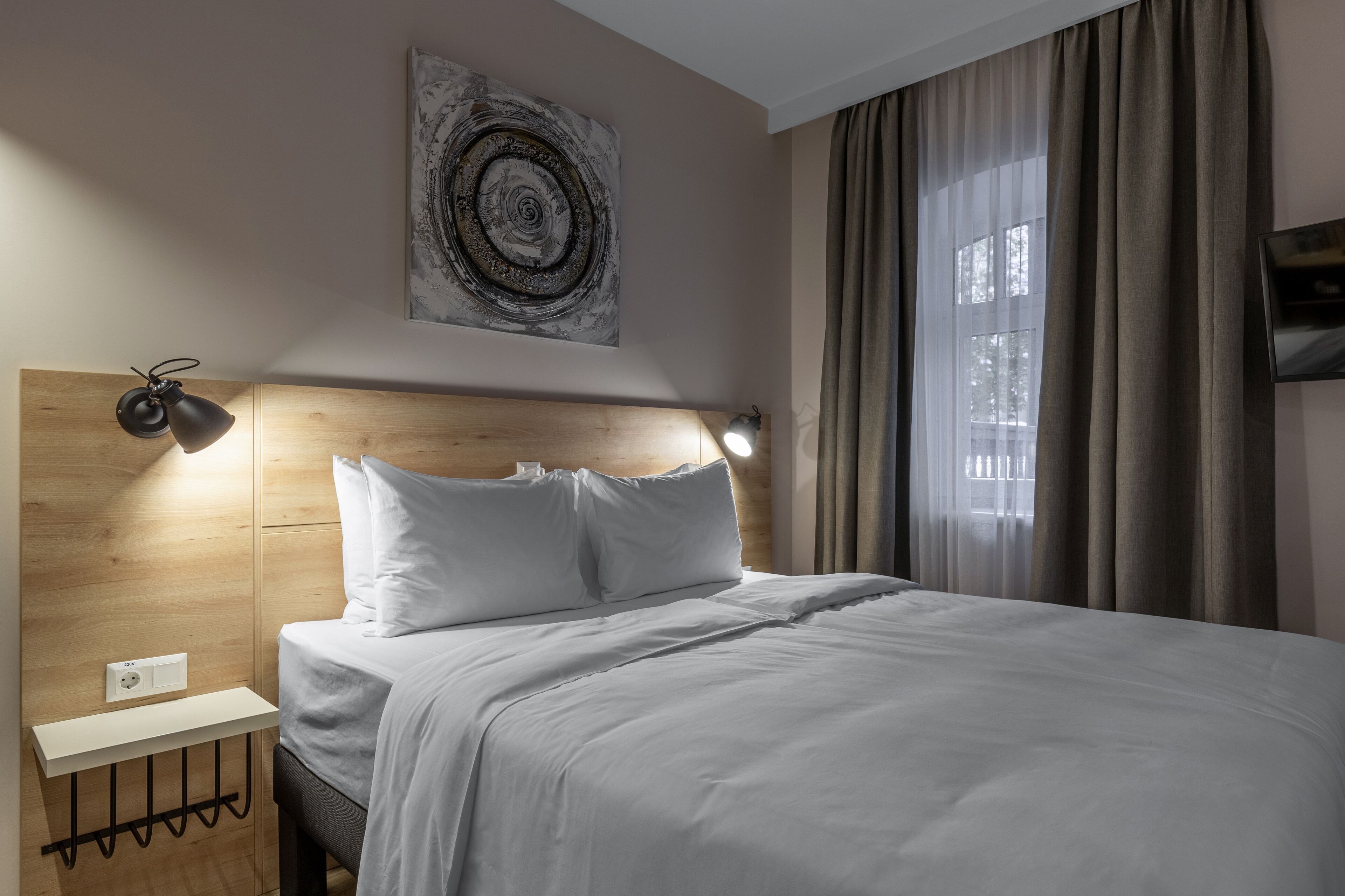 A modern hotel room with a wooden headboard, a bedside table with a lamp, a flat-screen TV, and a large window with curtains.