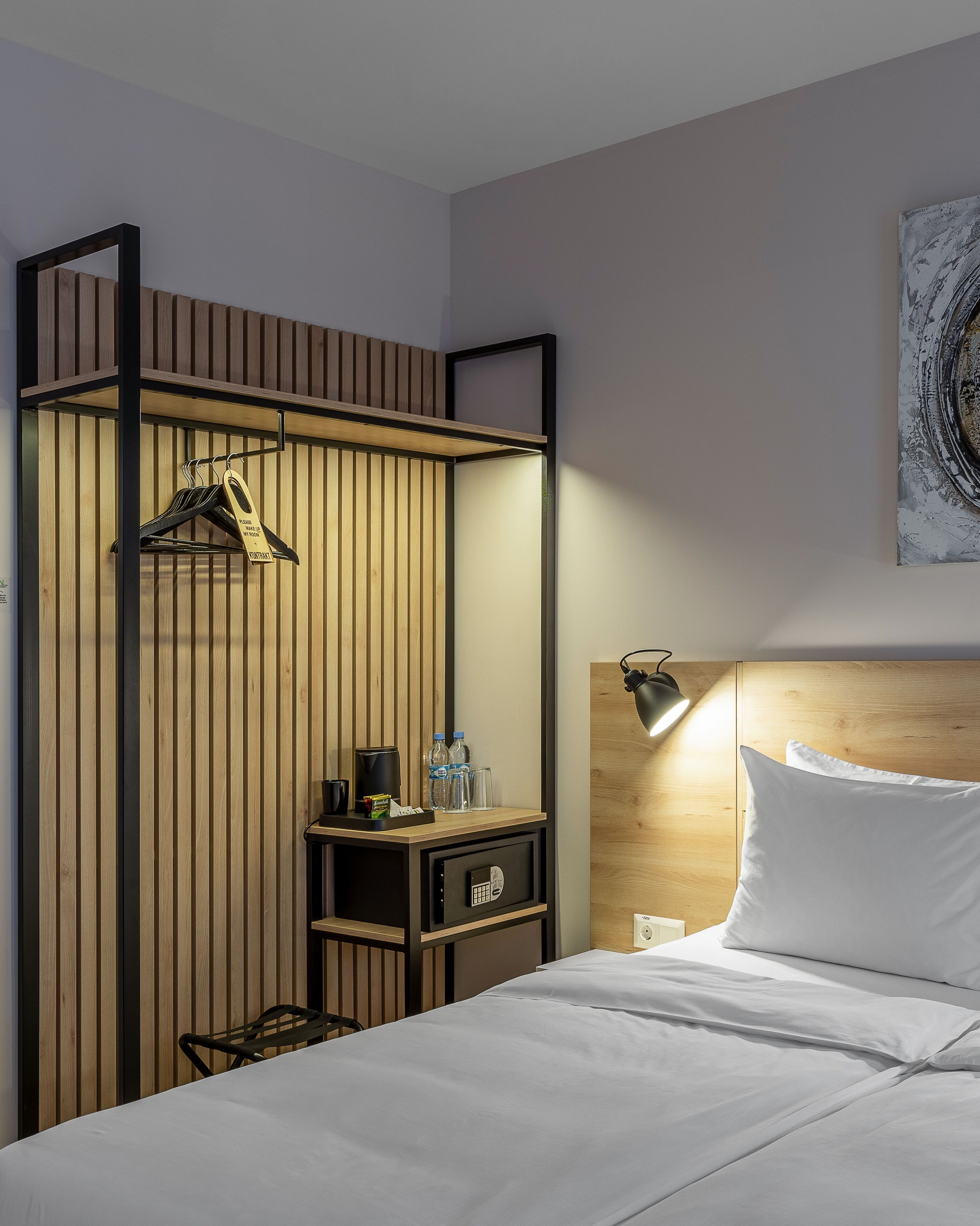 Una habitación de hotel con una cama, un separador de madera con un perchero, una mesita de noche con una lámpara y un cuadro en la pared.