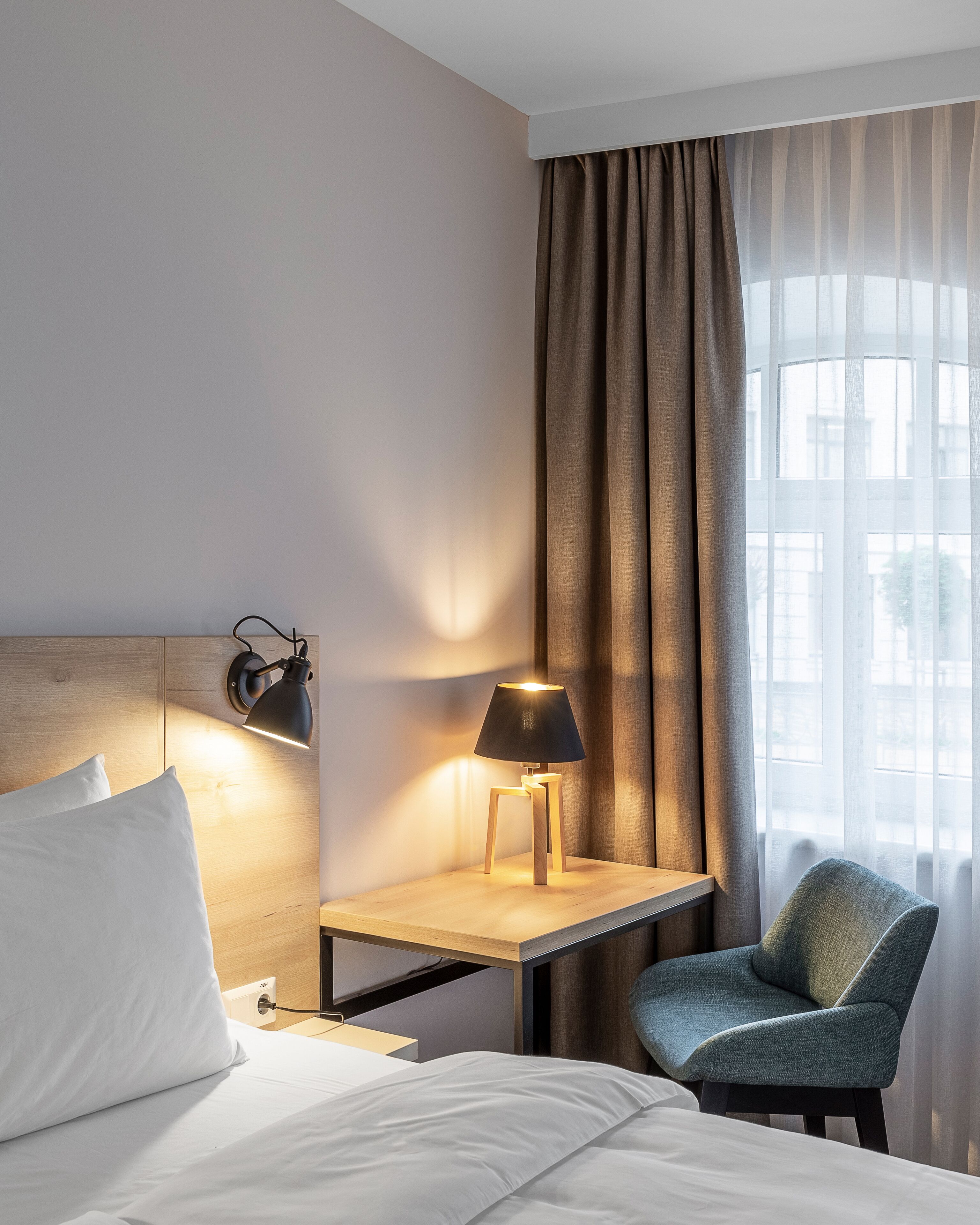 A hotel room with a bed, a desk with a lamp, a chair, and a window with curtains.