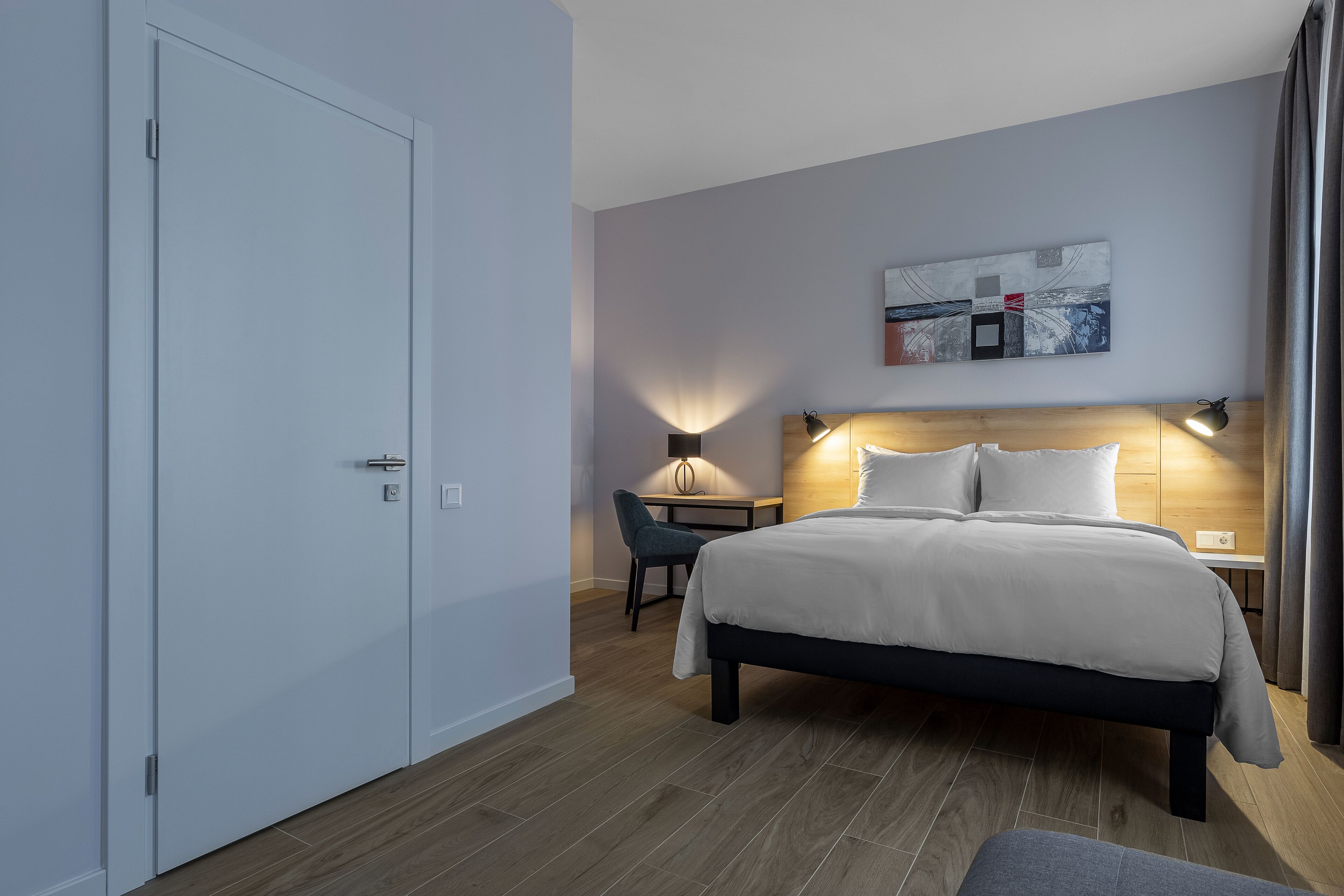 A modern hotel room with a large bed, a desk, and a chair. There is a painting on the wall and a lamp on the desk.