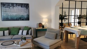 Un salon moderne avec un canapé, un fauteuil et une table basse, un coin repas avec une table dressée pour deux personnes, et un grand tableau abstrait accroché au mur.
