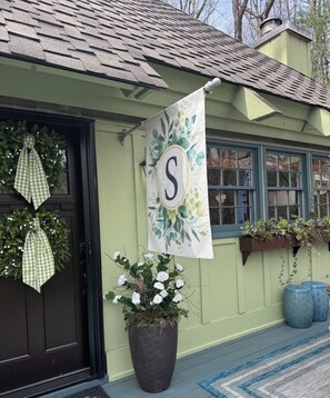 Una casa con puerta color marrón oscuro, fachada verde, una planta grande en maceta con flores blancas, un tapete de diseño azul y blanco y un banderín con una letra 'S' grande.
