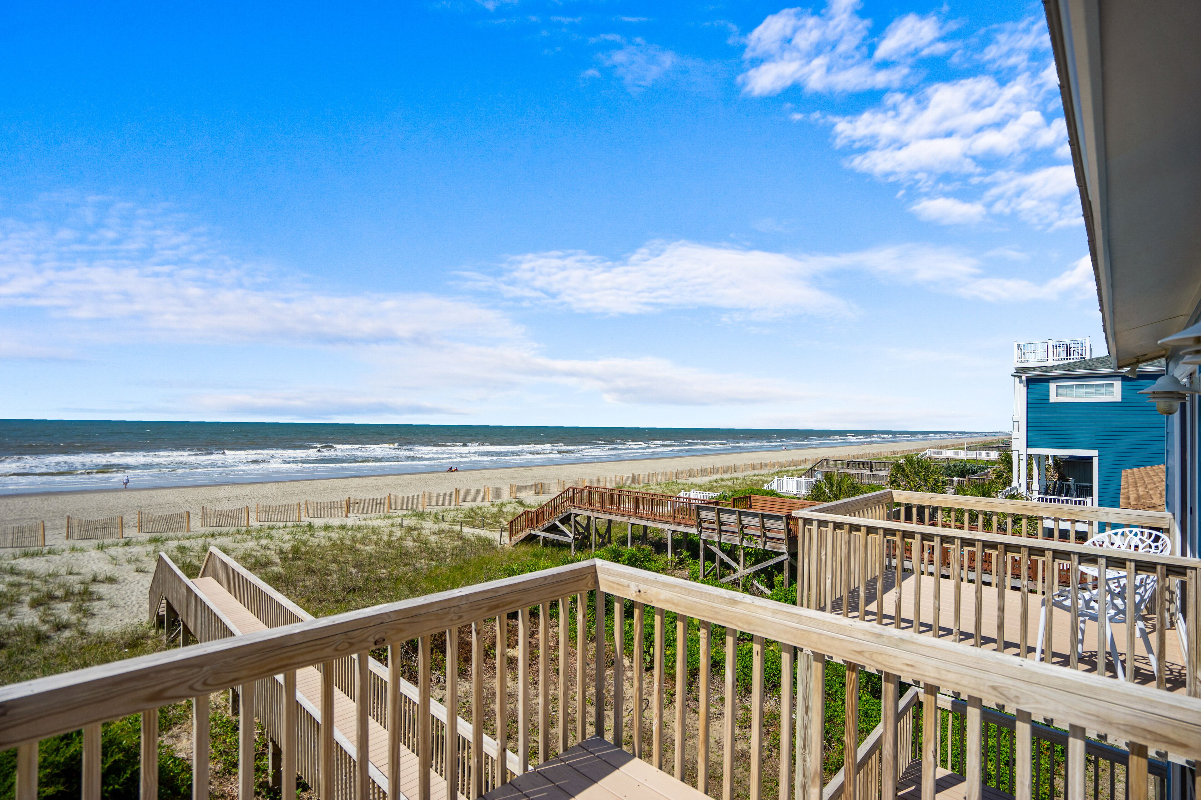A wooden deck overlooking a beach with a view of the ocean and a blue building with a balcony.
