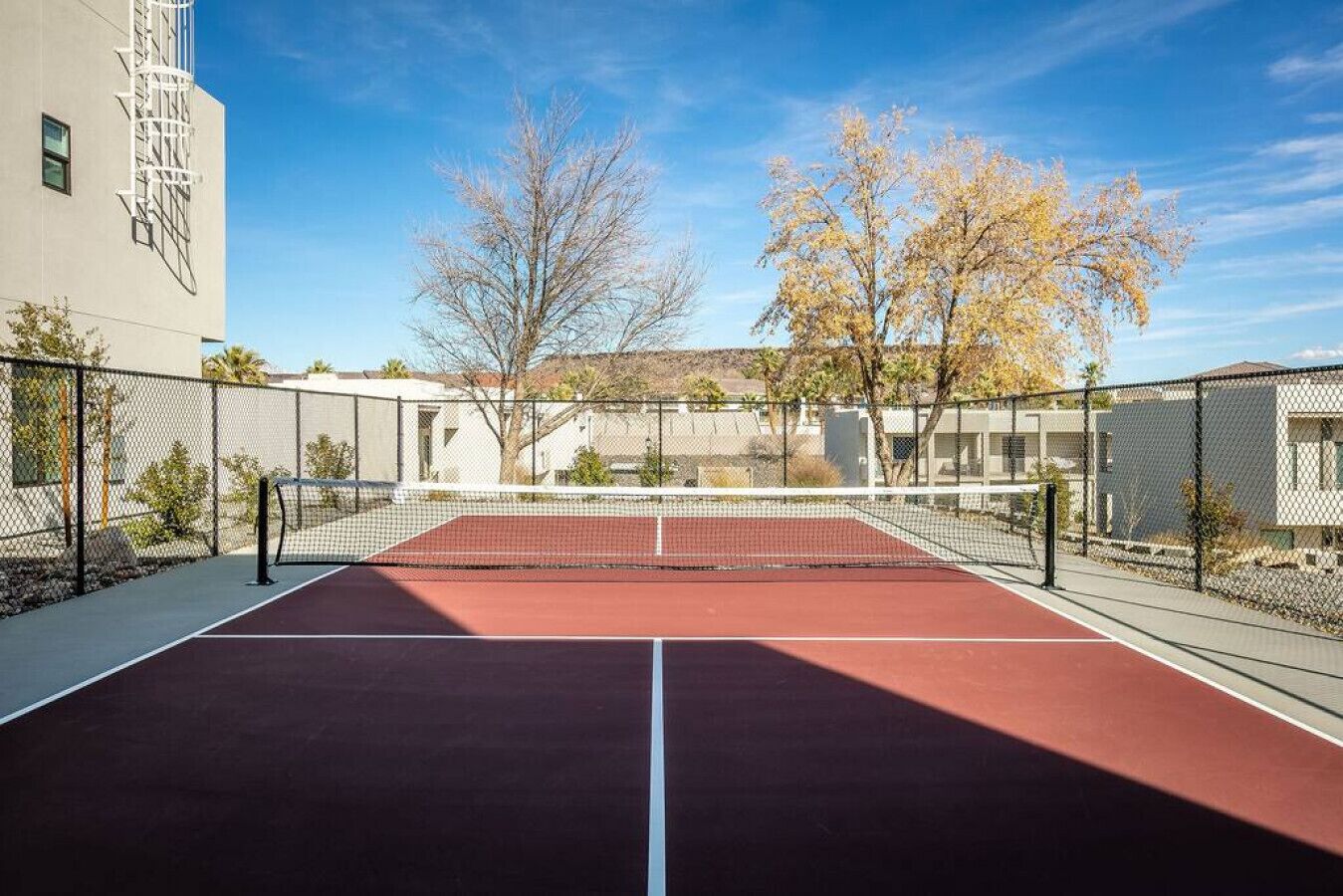 An outdoor tennis court with a red surface, surrounded by a chain-link fence, and set against a backdrop of trees and buildings.