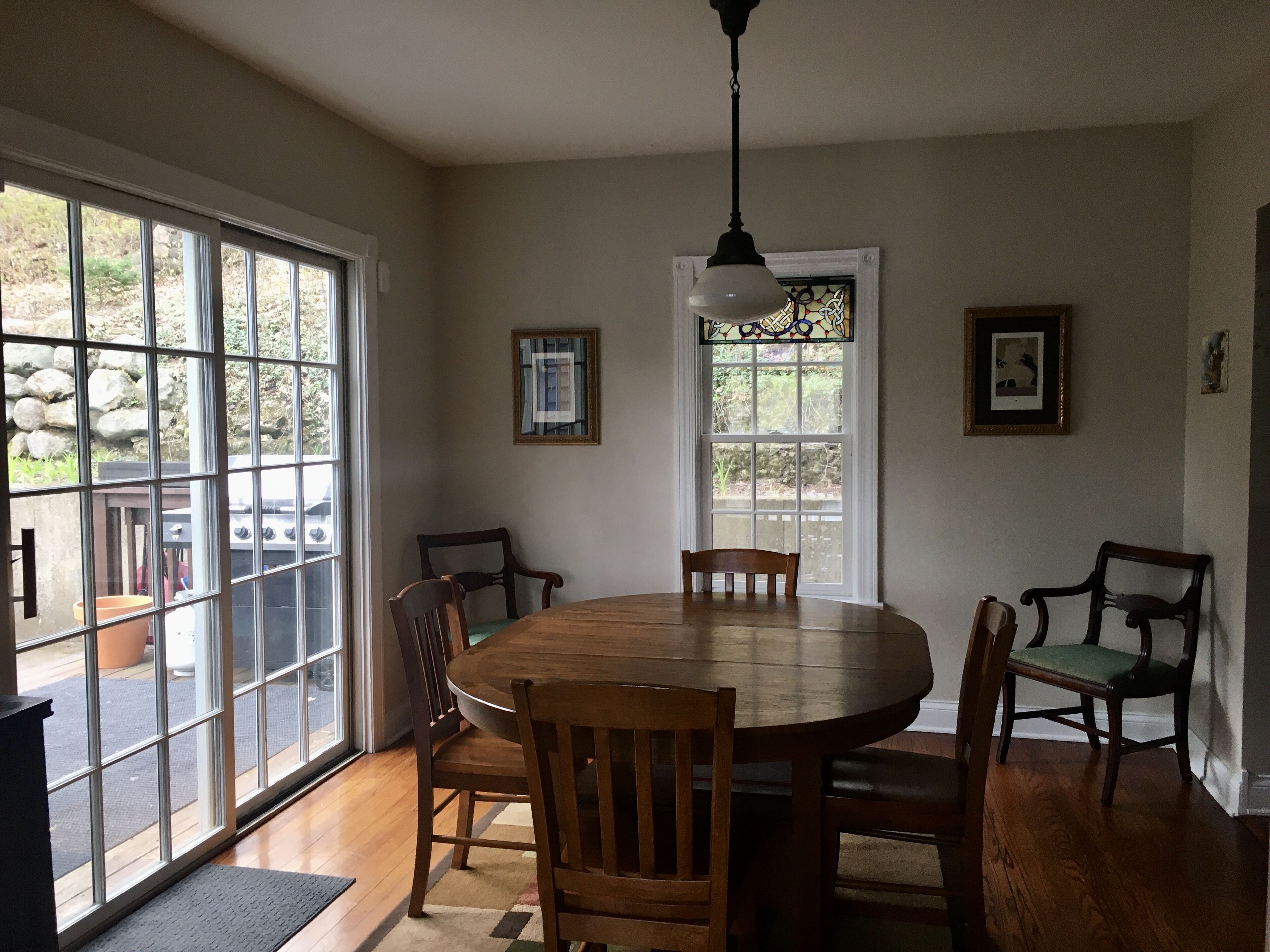 A dining area with a round wooden table and six chairs, large windows with a view of the outdoors, and a hanging light fixture.