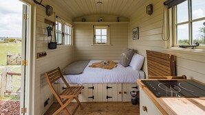 A cozy cabin with a bed, a wooden chair, a small window, and a stove. - Rumwold by Bloom Stays -  a rural shepherds hut sleeping 2 with amazing views! (Ashford)