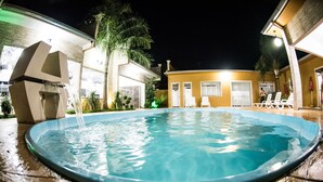 A swimming pool with a water feature, surrounded by a building with a curved roof and a palm tree. - Hotel Tito's (Capanema)
