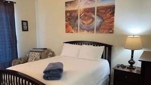 Una cama bien hecha con edredón blanco y almohadas, una lámpara de noche, una mesita de noche con lámpara y una obra de arte en la pared que representa un cañón.