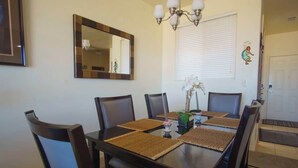 Un comedor con una mesa rectangular, seis sillas, una araña, un espejo y una ventana con persianas.