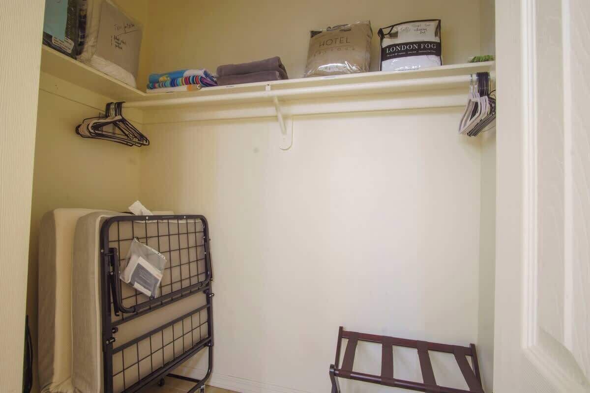 Habitación de hotel con cama, un pequeño banco de madera, un carro de lavandería metálico, un estante blanco con artículos de aseo y un perchero.