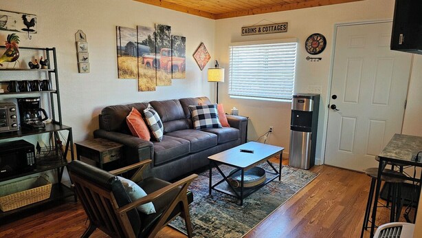 Una sala de estar acogedora con sofá, una mesa de centro y una mesa de comedor. Hay un refrigerador, un reloj y un cartel que indica 'Cabins & Cottages'.