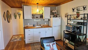 Cocina compacta con armarios de madera, un refrigerador, una cafetera y un comedor con una silla y una mesa.