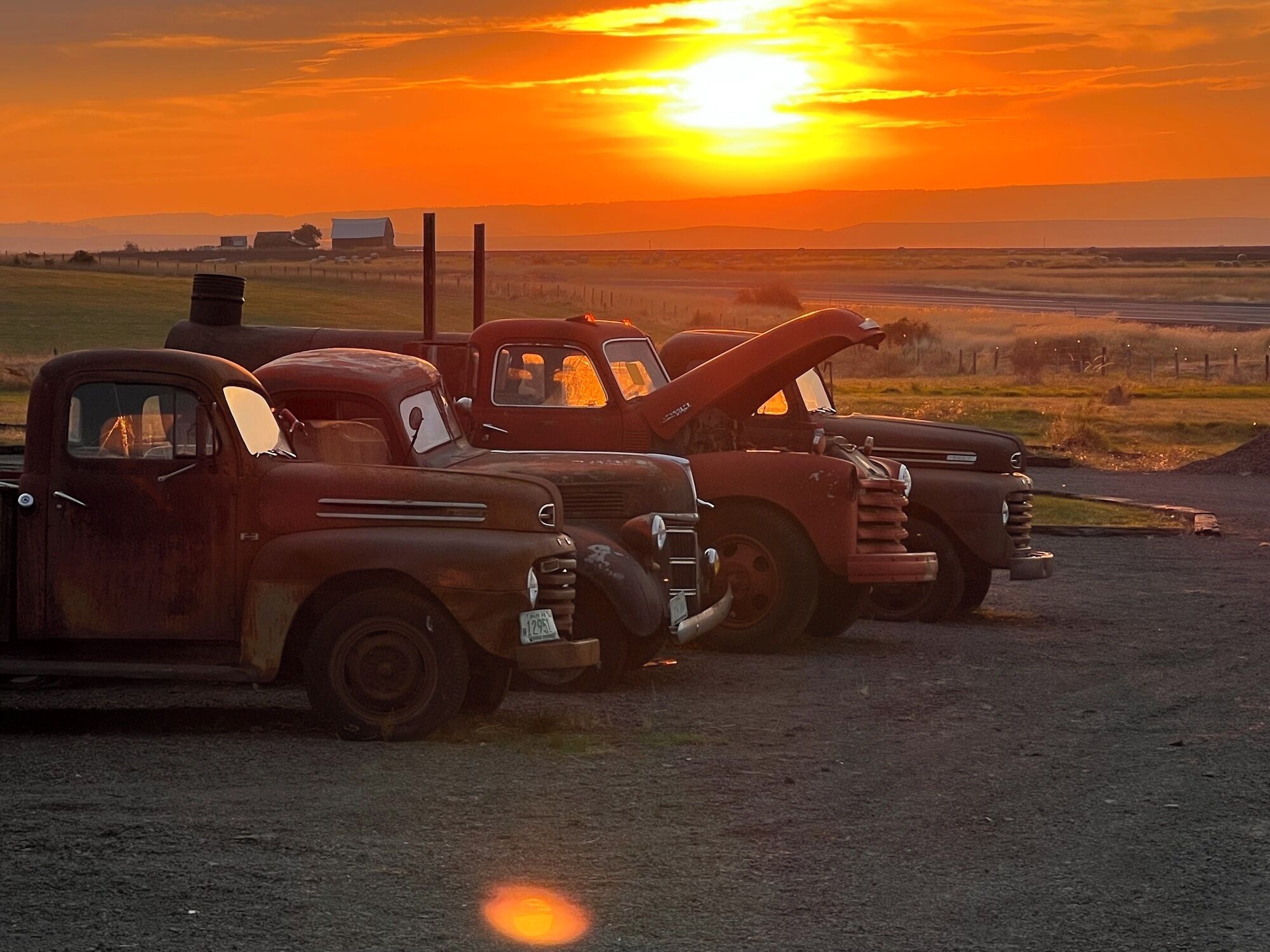 Tres camiones antiguos estacionados en un camino de tierra con un atardecer de fondo.