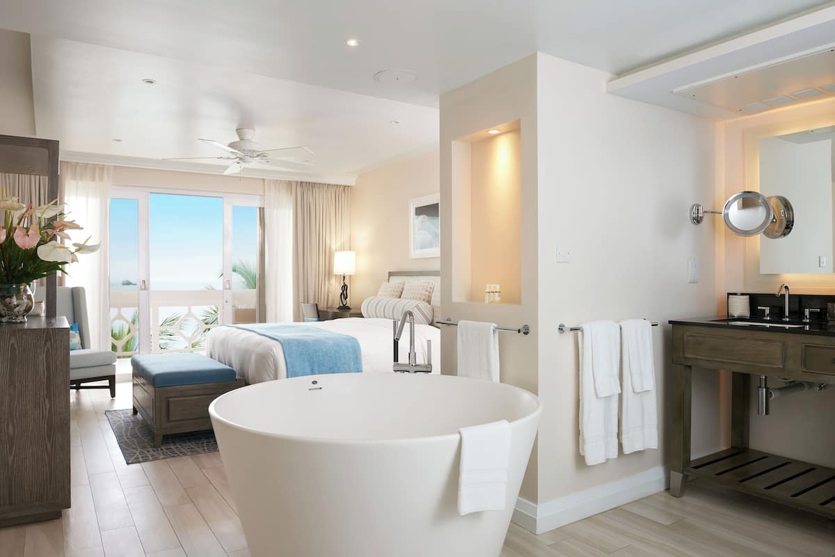A modern hotel room with a large bathtub, a bed with a blue throw, a desk with a lamp, and a view of the outdoors.