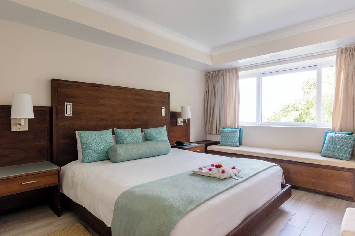 A hotel room with a large bed, bedside tables, a window with curtains, and a built-in bench with cushions.