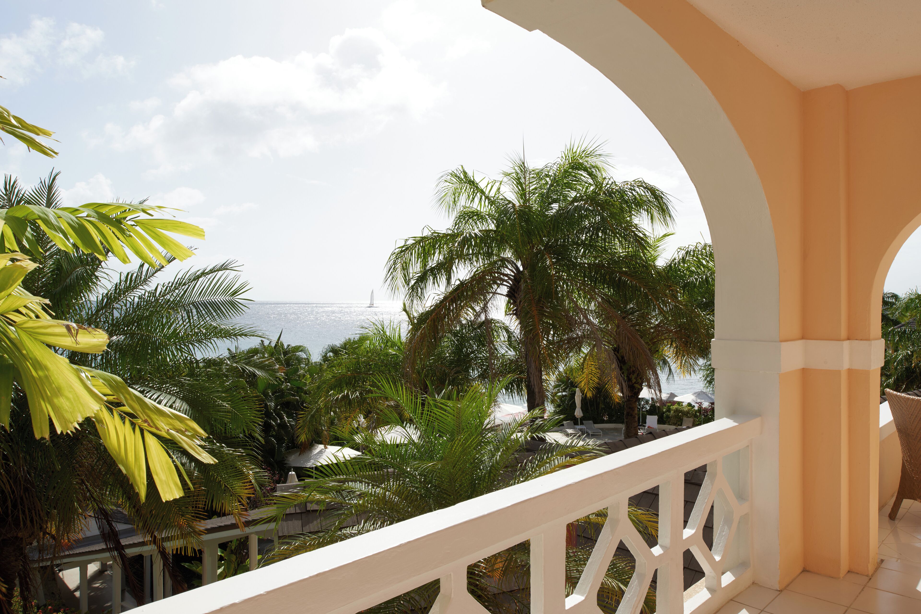 A balcony with a white railing, overlooking a beach with palm trees and a view of the ocean.