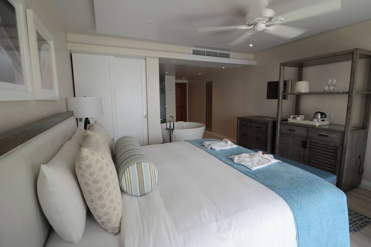 A hotel room with a large bed, a ceiling fan, a bathroom with a sink and mirror, and a wardrobe with a mirror.