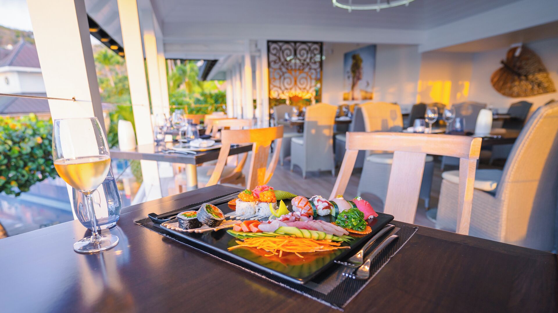 Une table à manger avec un plateau de sushis, un verre de vin blanc et une vue sur l’intérieur d’un restaurant, avec ses chaises et ses tables.
