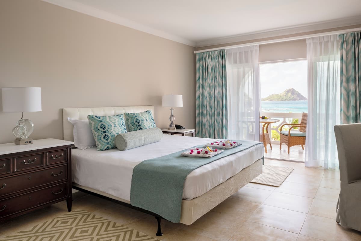 A hotel room with a bed, two bedside tables, a dresser, and a balcony with a view of the sea.