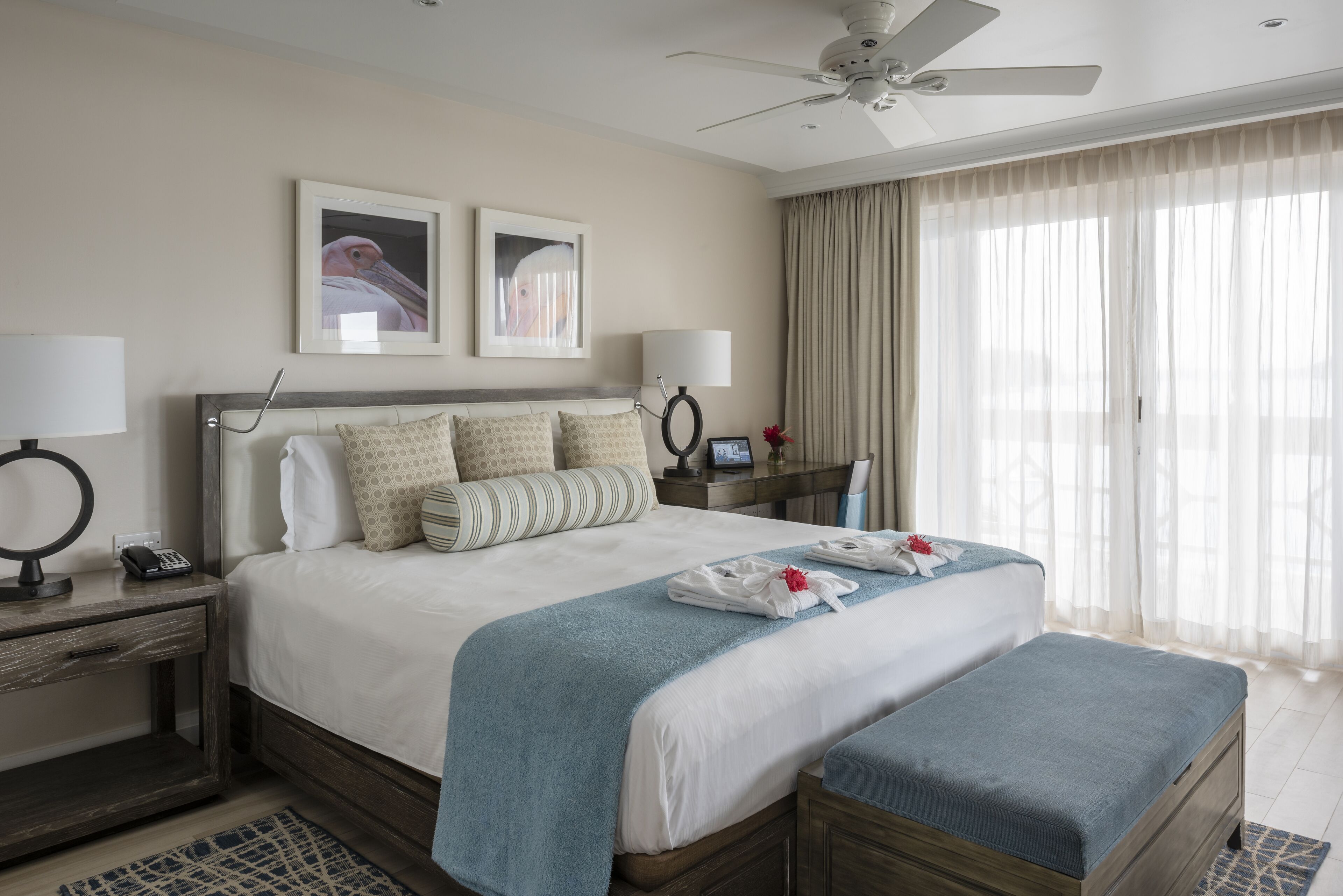 A hotel room with a large bed, a bedside table with a lamp, a desk with a chair, and a large window with curtains.