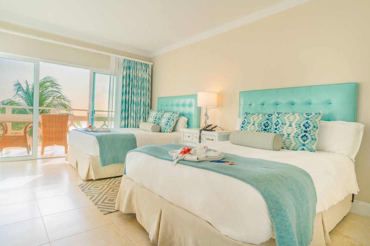 A hotel room with two beds, a balcony with a wicker chair, and a view of palm trees.