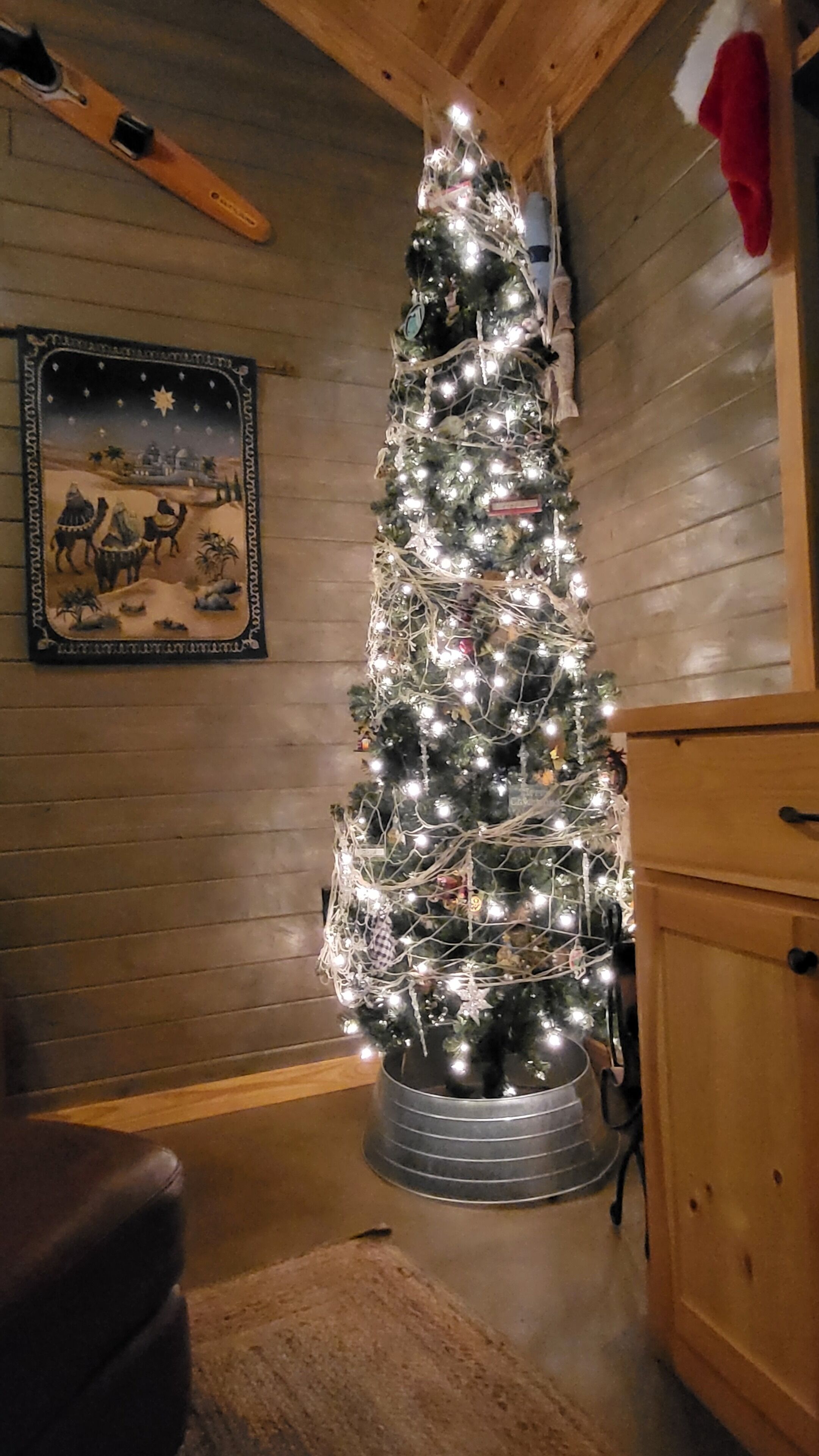 A Christmas tree with lights in a corner of a room with wooden walls and a wooden cabinet.