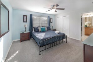 Ein Schlafzimmer mit einem Bett, Nachttischen, einem Deckenventilator, einem Fenster mit Vorhängen und einer Tür zu einem Badezimmer.