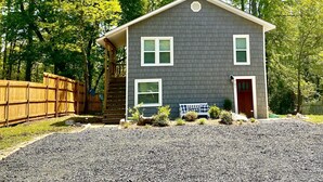 A two-story house with a dark gray exterior, white trim, and a red front door. There is a wooden fence to the left and a bench with plants in front of the house.
