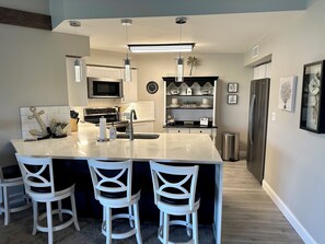 Una cocina moderna con una isla central, taburetes blancos de bar, un microondas, electrodomésticos de acero inoxidable y una repisa decorativa con elementos de decoración.