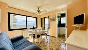 A living room with a sofa, a dining table with chairs, a ceiling fan, and a view through large windows.